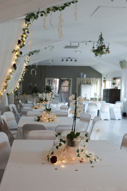 An elegantly decorated banquet hall with soft ambient lighting and white draped ceiling, featuring floral arrangements placed on tables. The tables are adorned with small vases holding white orchids a