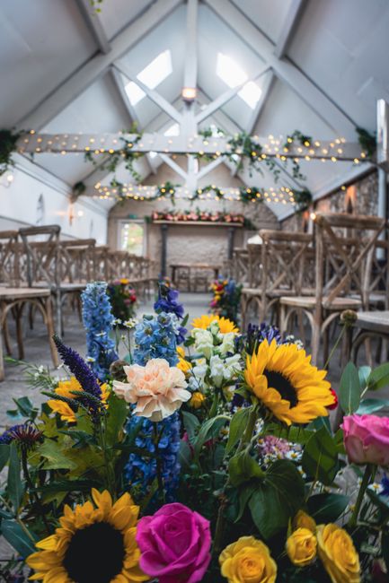 A vibrant floral arrangement featuring bright yellow sunflowers, pink roses, blue delphiniums, white lisianthus, and purple veronica stems, displayed in a lush bouquet with green foliage. The arrangem