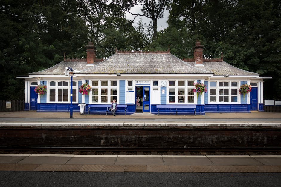 A charming, vintage-style train station building painted in blue and white, situated at the Coulsdon Town station area. The single-story structure features large sash windows decorated with vibrant ha