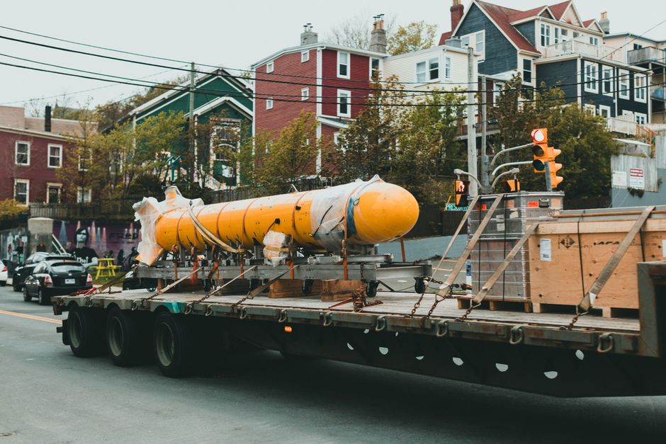 The image shows a large yellow cylindrical object, possibly a submarine or industrial component, loaded onto a flatbed truck for transportation. The object is secured with straps and surrounded by woo