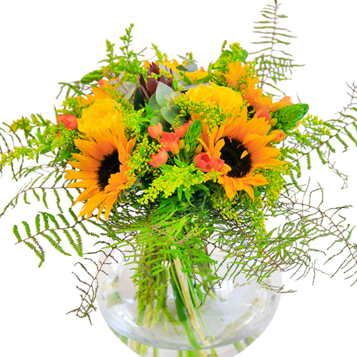 The first impression given by this Coulsdon bouquet is one of radiant, sunlit abundance-a scene where luminous yellow and orange sunflowers mingle with plush garden roses, each bloom unfurling in layers of lively warmth. The powerful energy of the sunflowers, their dark centers and surrounding petals like miniature sunbursts, is balanced by the soft, intricate folds of the roses, rich in both texture and fragrance. Spotted throughout, clusters of shiny red Hypericum berries create vivid focal points, standing out against the generous green backdrop provided by feather-light ferns and airy sprays of goldenrod. Slender flower spikes and soft eucalyptus leaves add further dimension, their bluish-green tones evoking the peaceful green belts that border the area around Fox Lane. The arrangement is artfully placed in a round, clear glass vase, where the submerged stems appear to twist and dance in crystal water, the whole piece illuminated by soft natural light. This bouquet encapsulates the heart and warmth of Coulsdon itself: perfect for brightening an anniversary, celebrating a milestone, or welcoming guests to a sunlit home, all while reflecting the lush, serene beauty of the neighbourhood.