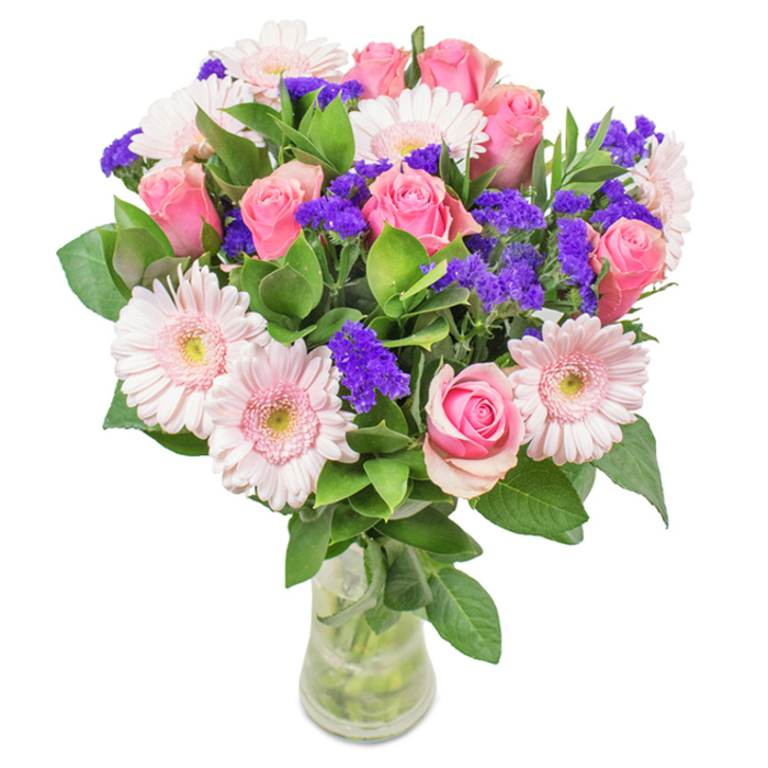 An artfully arranged bouquet presented in a clear glass vase stands out against a clean white background, echoing the natural light of Coulsdon's leafy neighborhoods. Central to the display are 7-9 pink roses, some elegantly unfurling while others remain tightly spiraled, emphasizing classic beauty and layered tints. Encircling these are 5-7 pale pink gerbera daisies, recognized by their generous, round heads and subtly lighter centers. Amidst the pastels, clusters of bright purple statice provide vibrant contrast and tactile dimension. The abundant green foliage creates a lush underlayer, enhancing the freshness and vibrancy one might find on a walk near Rickman Hill Park. The glass vase reveals the stems and clean water, reinforcing a sense of natural abundance and purity. This bouquet-well-suited to celebrate birthdays, anniversaries, or to brighten an office corner-embodies joy, affection, and a little of that local Coulsdon charm, making it a much-loved gift from Coulsdon Flowers.